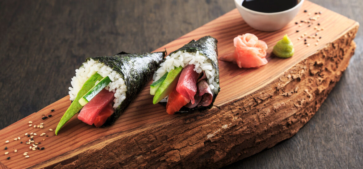 un sushi temaki entre 2 palitos para comer. de fondo un plato de sushi variado